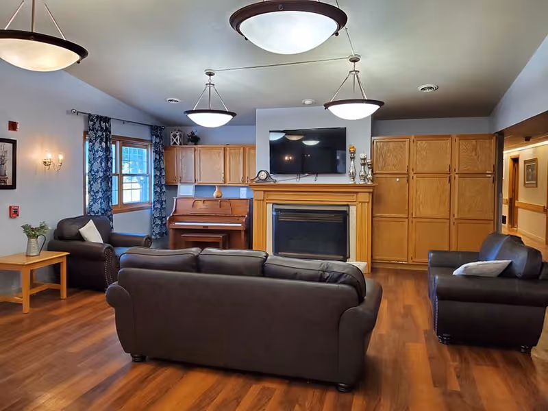 A cozy senior living common area with wooden flooring, a dark brown leather sofa and armchairs, a wooden piano, a fireplace with a mounted flat-screen TV above it, wooden cabinets, and windows with blue patterned curtains.