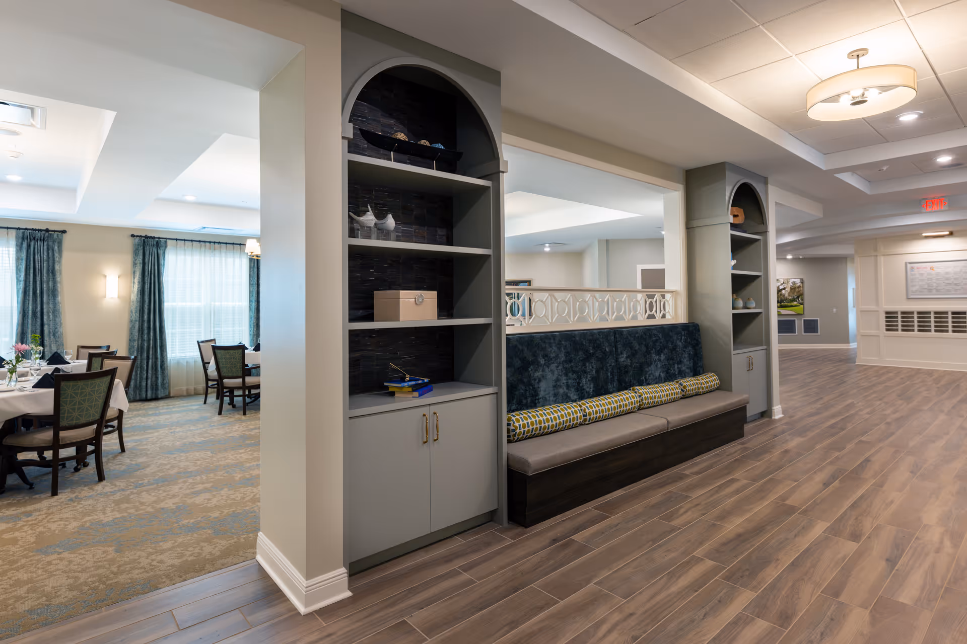 Interior view of a senior living facility showing a hallway with wood flooring and built-in shelving units with decorative items. To the left, there is a dining area with tables and chairs, set with white tablecloths and floral centerpieces. The space is well-lit with ceiling lights and natural light from windows with blue curtains.