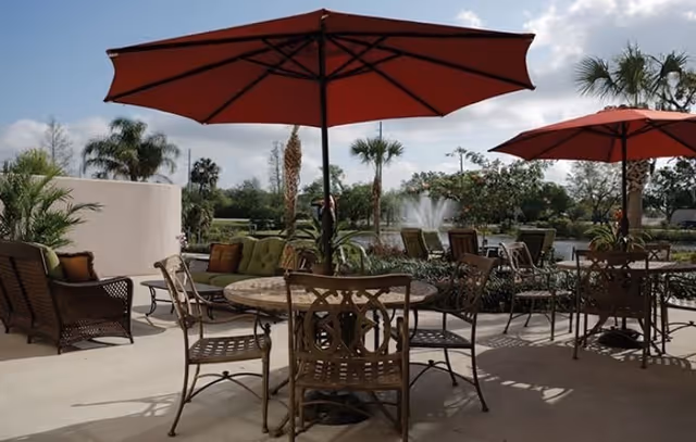 Outdoor patio area with metal tables and chairs under large red umbrellas, surrounded by palm trees and greenery, with a water fountain visible in the background.