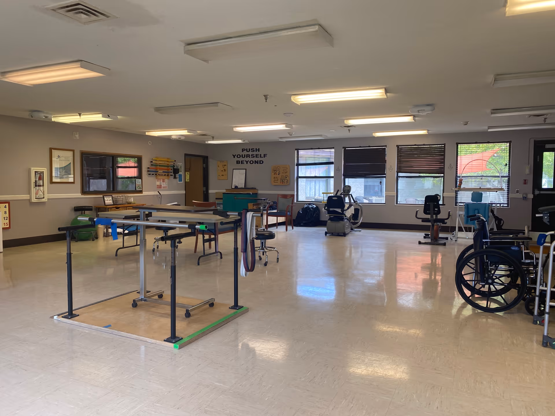 Spacious therapy/exercise room with parallel bars, exercise machines and wheelchairs under fluorescent lights