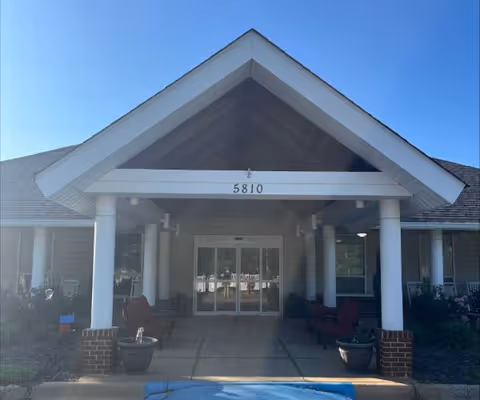 Entrance of a building with a covered porch supported by white columns and a peaked roof. The building number 5810 is displayed above the entrance doors. There are potted plants and outdoor seating on either side of the walkway leading to the glass double doors.