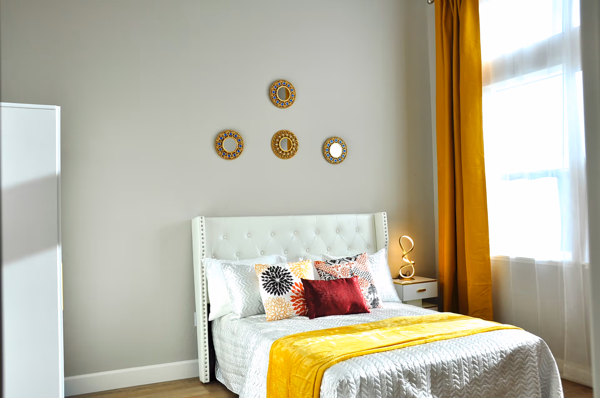 Bright bedroom with a white tufted bed, colorful pillows, a yellow throw and curtains, and decorative round mirrors on the wall.
