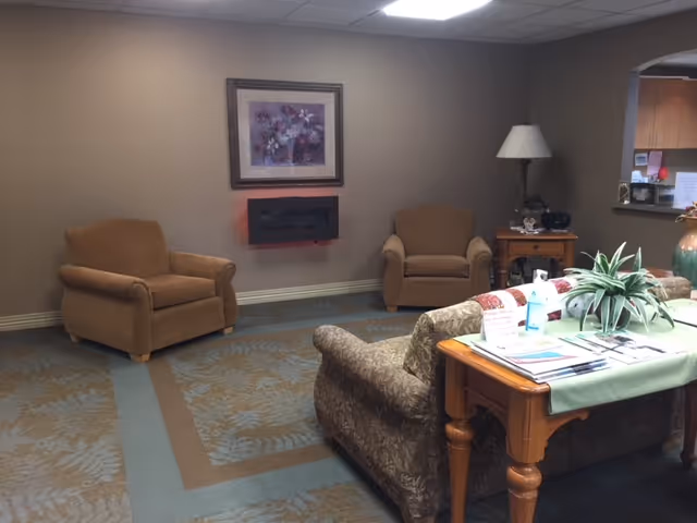A cozy living room area with two brown armchairs against a beige wall, a framed floral painting above a modern wall-mounted electric fireplace, a patterned sofa, a wooden side table with a lamp and decorative items, and a wooden table with a green table runner and various papers and a plant on it.