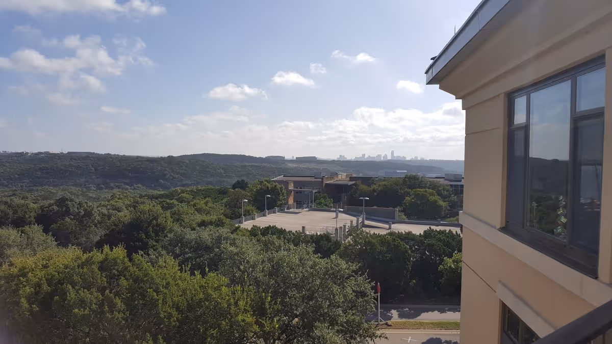 View from a building showing treetops and hills with a distant city skyline and part of the building facade on the right.