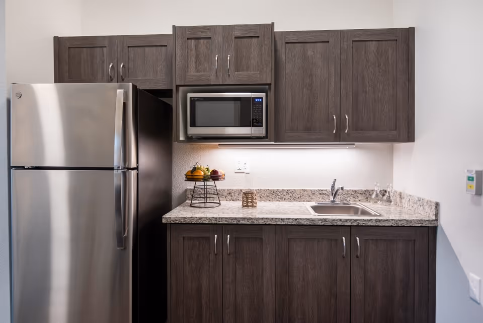 A modern kitchenette featuring dark wood cabinets, a stainless steel refrigerator, a microwave mounted above the countertop, a small sink, and a fruit basket on the granite countertop.