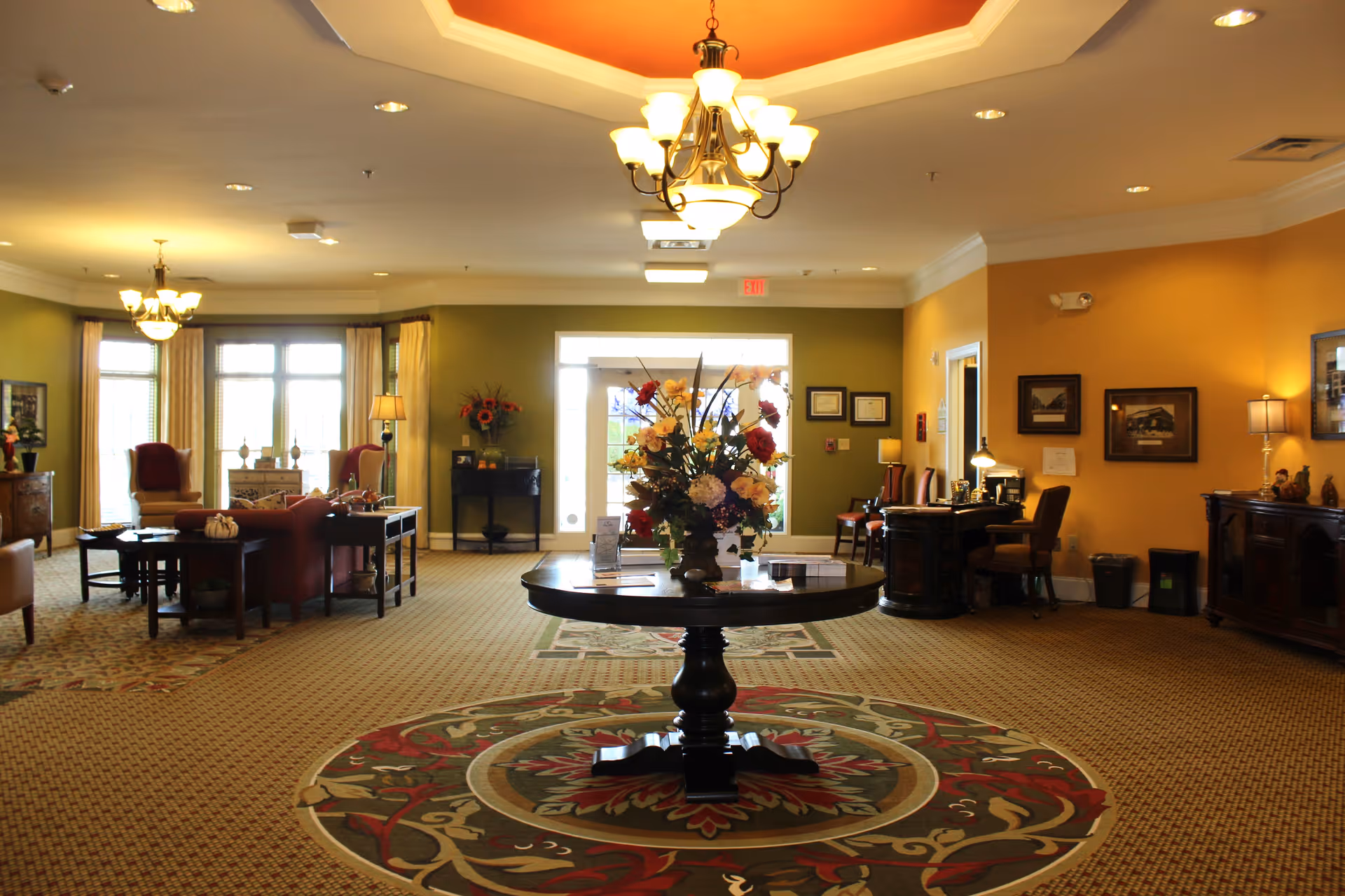 A spacious senior living facility common area with warm yellow and green walls, a round table with a large floral arrangement in the center, comfortable seating including armchairs and sofas, decorative lamps, framed pictures on the walls, and a patterned carpet with a circular design under the table.