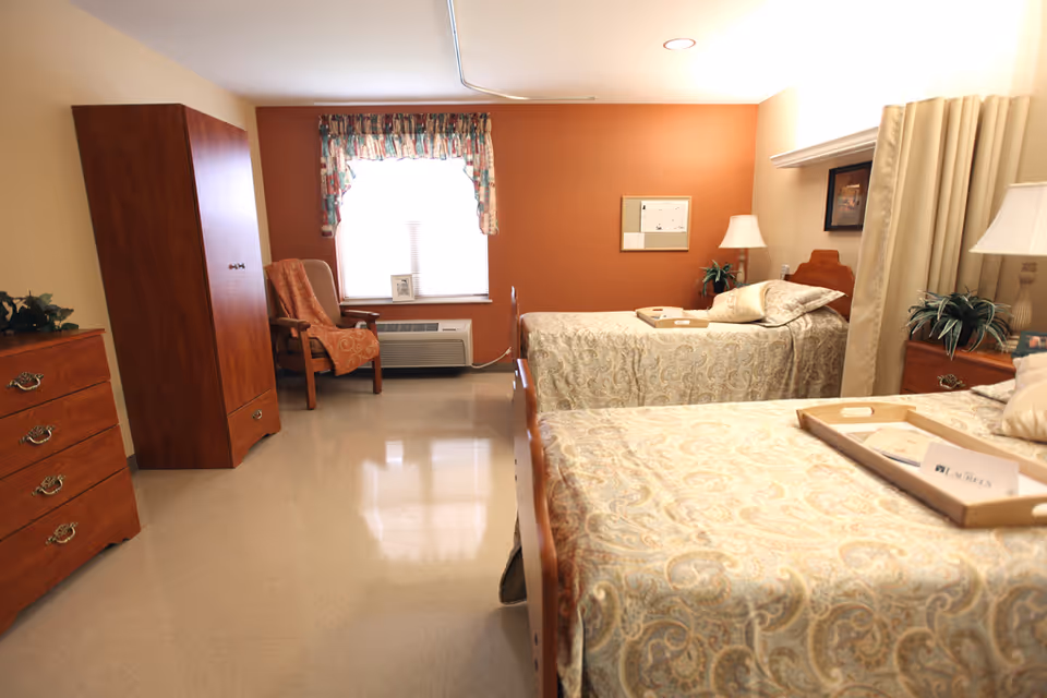 A senior living facility bedroom with two twin beds covered in patterned bedspreads. Each bed has a wooden headboard and a tray on top. There is a wooden dresser and wardrobe on the left side of the room, and an upholstered armchair near a window with floral curtains. The walls are painted beige and one accent wall is a warm orange color. Two table lamps and some plants add to the cozy atmosphere.