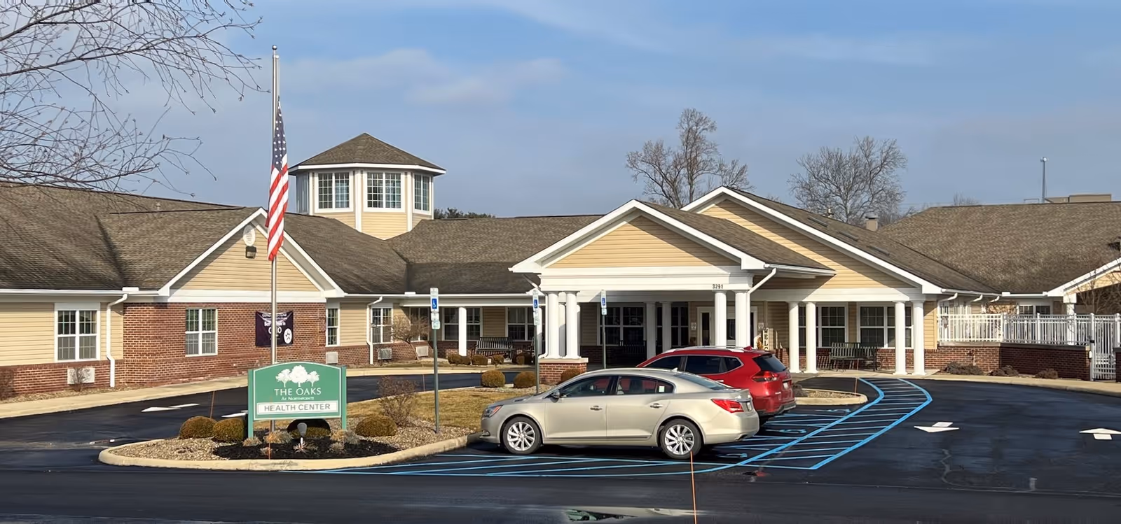 Exterior view of The Oaks Rehabilitation & Healthcare Center LLC building with a beige and brick facade, a covered entrance supported by white columns, an American flag on a flagpole, and two cars parked in front in designated parking spaces with blue markings.