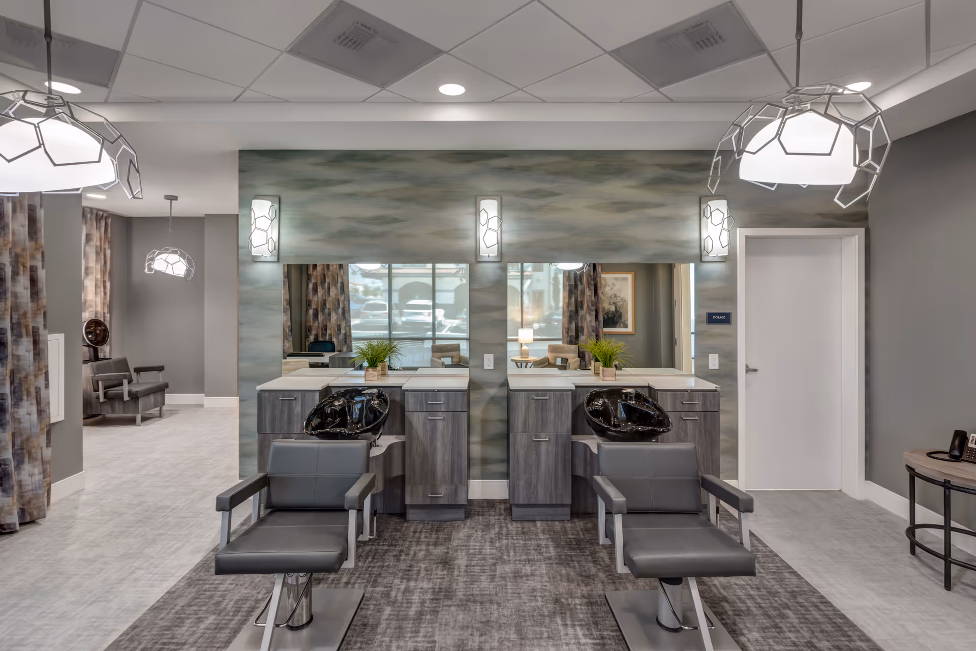 Modern salon area with two styling chairs, shampoo sinks, and mirrors in a senior living facility interior.