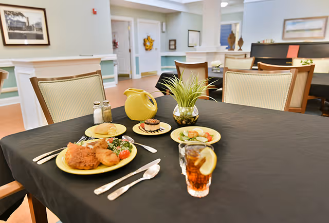 A communal dining table set with plates of food, a glass of iced tea with lemon, a small plant centerpiece and chairs in a dining room.