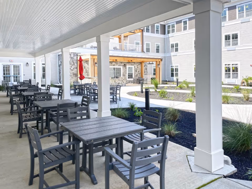Covered outdoor patio area with multiple black tables and chairs arranged for seating. The patio overlooks a landscaped courtyard with plants and a stone building facade in the background. A red umbrella is closed and positioned near one of the tables.