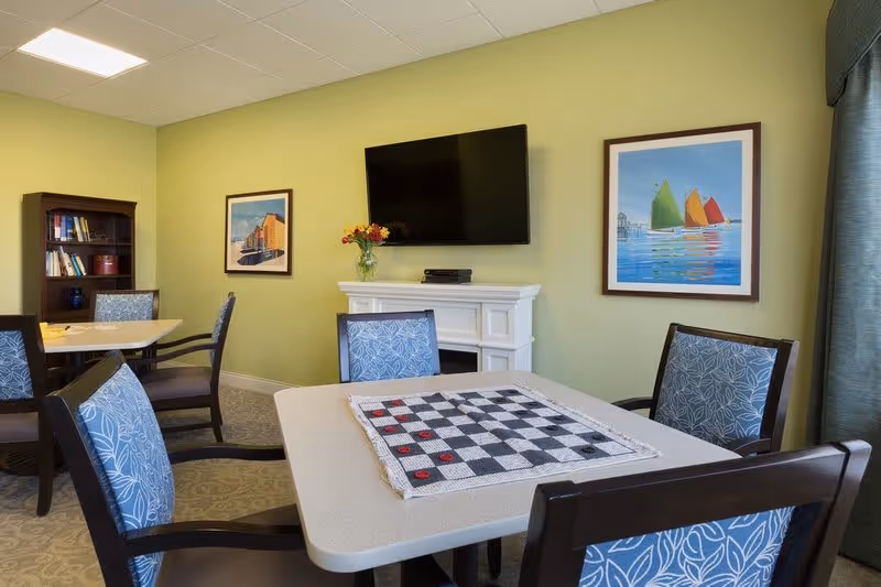 A cozy room with a table set up for a checkers game, surrounded by four chairs with blue floral upholstery. The room has light green walls, a mounted flat-screen TV above a white mantel, two framed paintings on the walls, a bookshelf with books, and a window with blue curtains.
