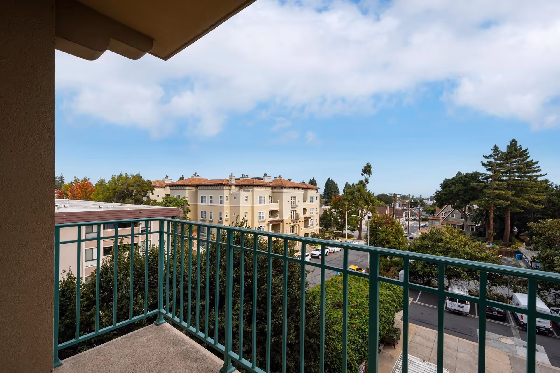 View from a balcony with green metal railings overlooking a street, trees, and a beige multi-story building with a red-tiled roof under a partly cloudy blue sky.