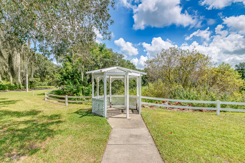 A small white gazebo with lattice sides situated at the end of a concrete pathway in a grassy outdoor area. Surrounding the gazebo are trees, bushes, and a white wooden fence under a partly cloudy blue sky.