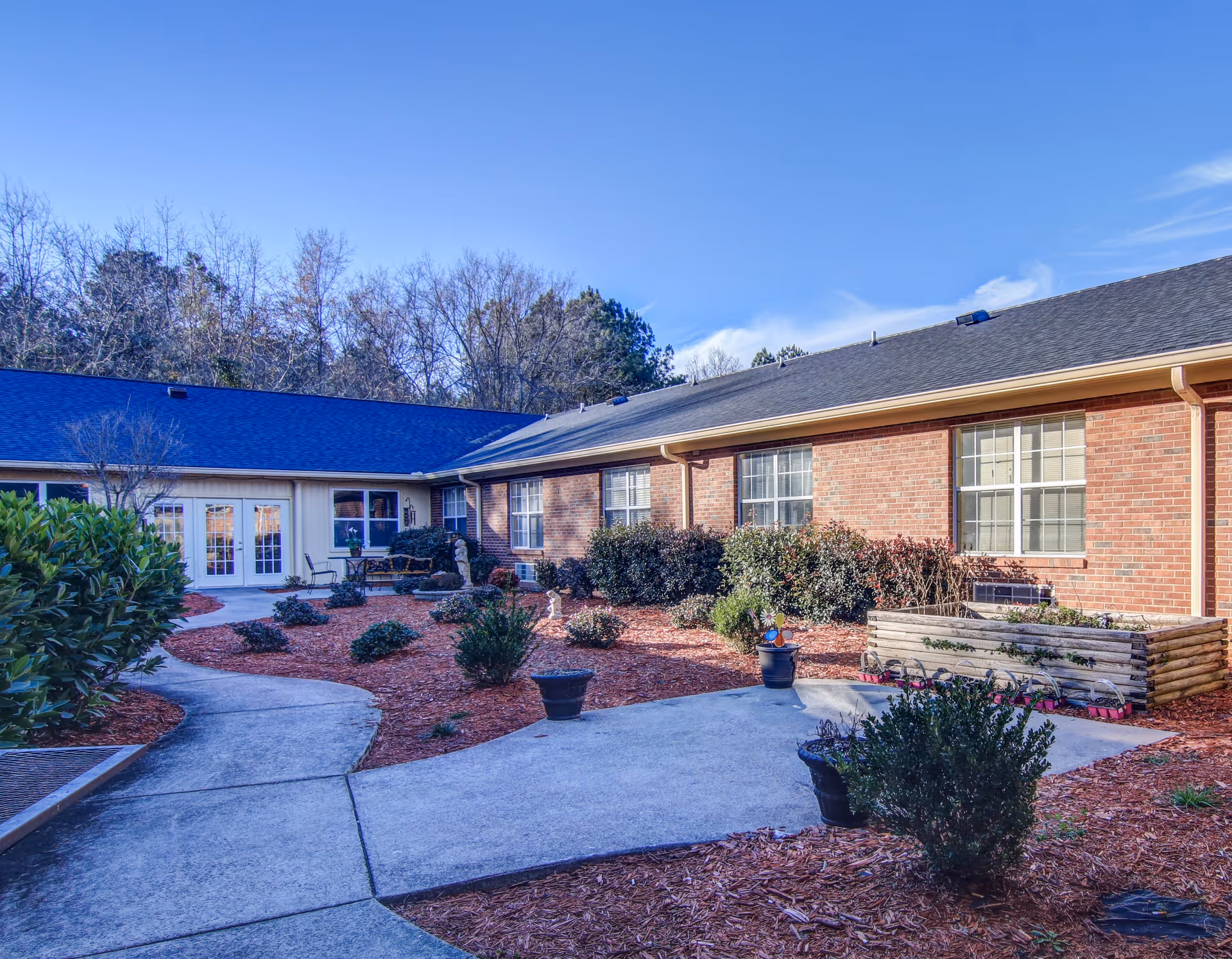 Outdoor courtyard area of a senior living facility with a concrete pathway winding through landscaped garden beds with bushes and potted plants. The building has brick walls and multiple windows, with a clear blue sky overhead.