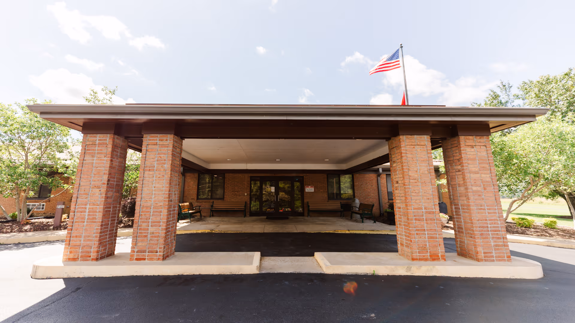 Front entrance of Laughlin Healthcare Center featuring a covered drop-off area supported by brick columns, with benches on either side and an American flag flying above the roof. Trees and greenery surround the building under a partly cloudy sky.