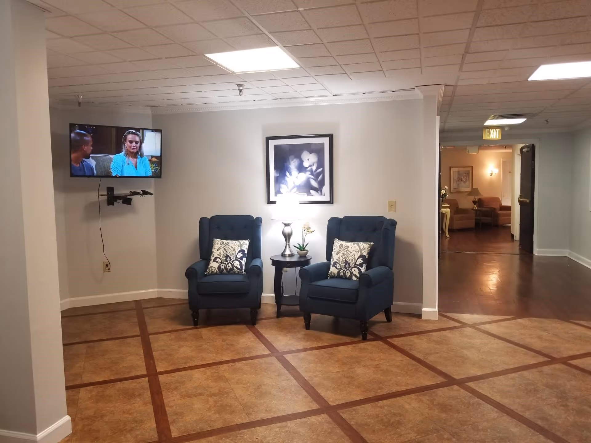 A cozy seating area in a senior living facility with two blue armchairs featuring patterned cushions, a small round table with a lamp and a small plant between them, a framed floral picture on the wall, and a wall-mounted TV showing a scene with two people. The floor has a tile pattern with brown borders, and there is a hallway leading to another room with more seating visible.