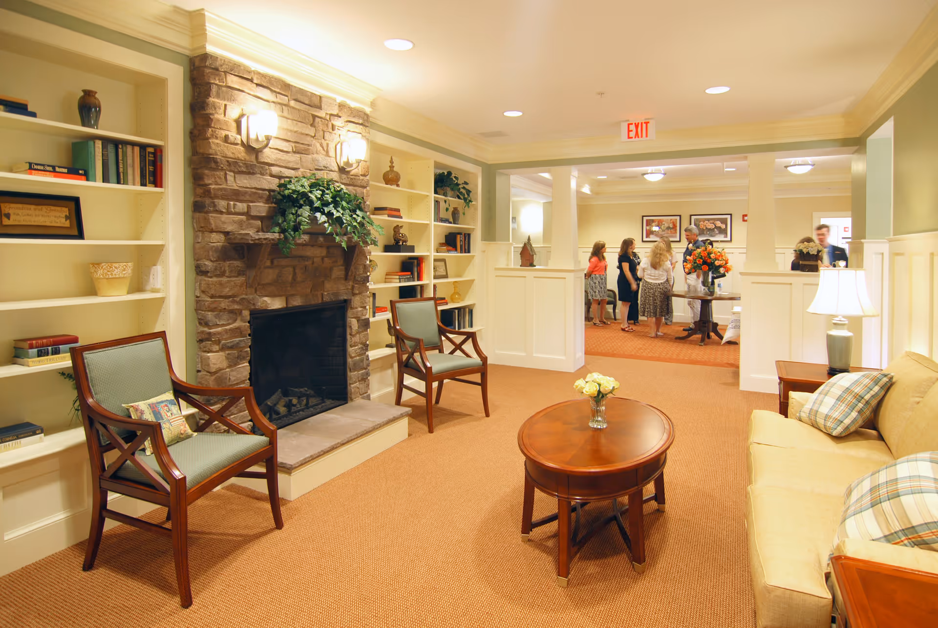 A cozy living room area in a senior living facility with a stone fireplace, two wooden chairs with green cushions, a wooden coffee table with a vase of flowers, a beige sofa with plaid pillows, built-in bookshelves with books and decorative items, and a group of people conversing in the background near an exit sign.