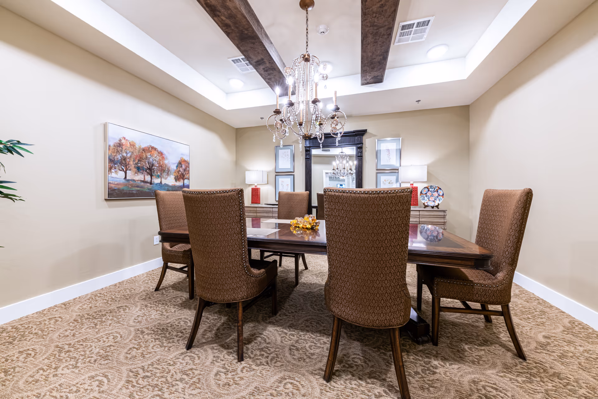 A formal dining room with a rectangular wooden table surrounded by six upholstered chairs. The room features a patterned carpet, beige walls, a chandelier hanging from a ceiling with wooden beams, a large mirror on the far wall, two table lamps with red bases, and a framed painting of trees on the left wall.