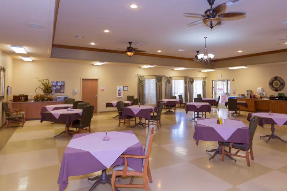 A spacious dining room with several round tables covered with purple tablecloths and light purple overlays. Each table is surrounded by chairs, and the room is well-lit with ceiling lights and ceiling fans. There are large windows with curtains along one wall and a counter area with various items on the right side of the room.