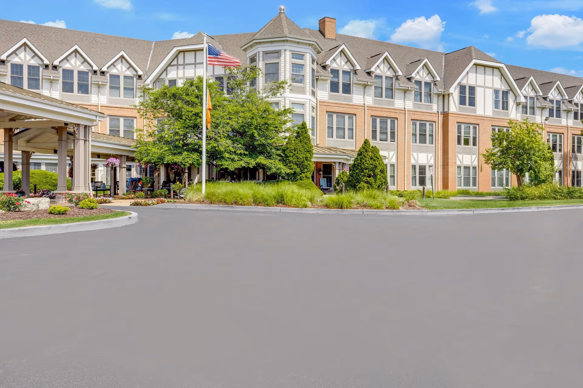 Exterior view of Sunrise of Des Peres senior living facility showing a large multi-story building with Tudor-style architecture, surrounded by well-maintained landscaping, trees, and a flagpole with an American flag in front under a blue sky with some clouds.