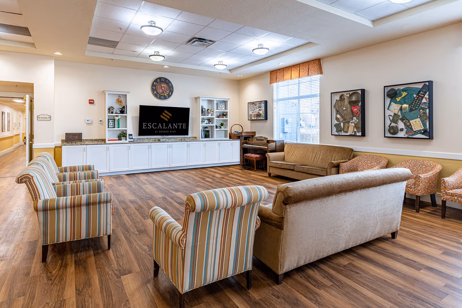 Spacious senior living lounge with sofas and armchairs arranged around a wall-mounted TV showing 'Escalante' and built-in cabinets.
