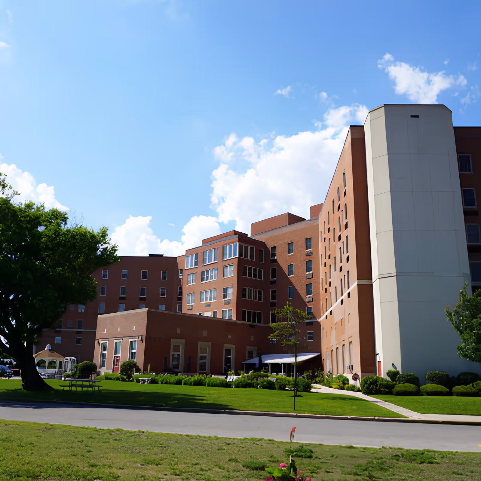 Exterior view of a multi-story brick building with large windows, surrounded by green lawns, trees, and a clear blue sky with some clouds.