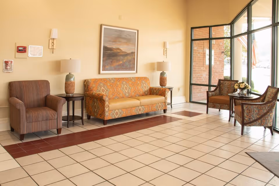 A seating area in a facility with a patterned orange sofa, two matching armchairs, and one striped armchair. There are two side tables with lamps on either side of the sofa, a small round table with a flower arrangement between the two armchairs near large windows, and a framed landscape painting on the wall. The floor is tiled with a pattern of white and reddish-brown tiles.