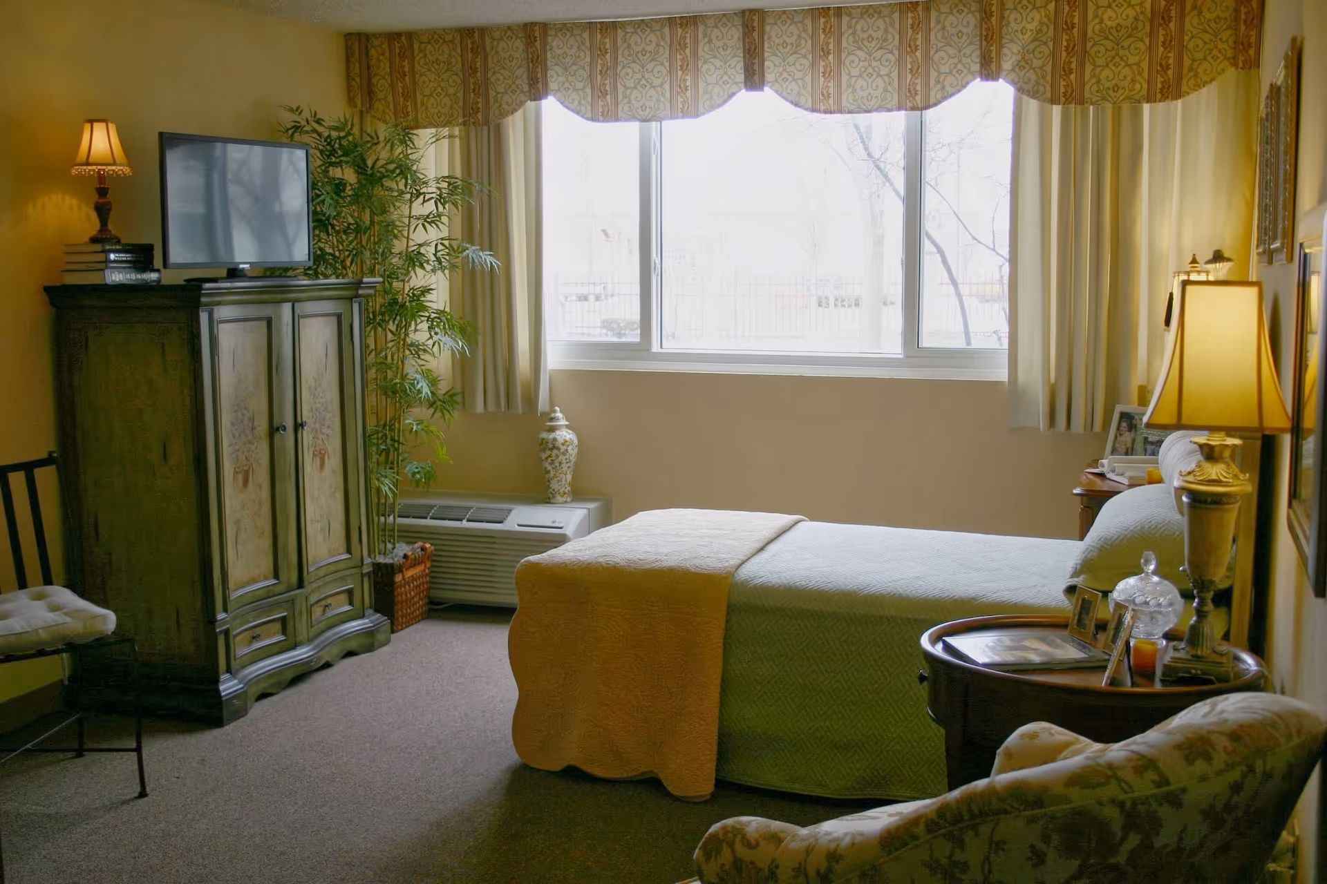 A cozy bedroom in a retirement community featuring a single bed with a yellow blanket and green bedspread, a wooden armoire with a TV on top, a potted plant, a window with curtains and valance, a side table with a lamp and framed photos, and a cushioned armchair.
