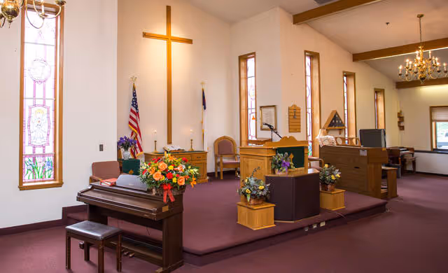 Interior of a chapel with a large wooden cross on the wall, stained glass windows, an American flag, a Christian flag, a piano with a floral arrangement, a lectern with a microphone, and several chairs arranged on a raised platform.