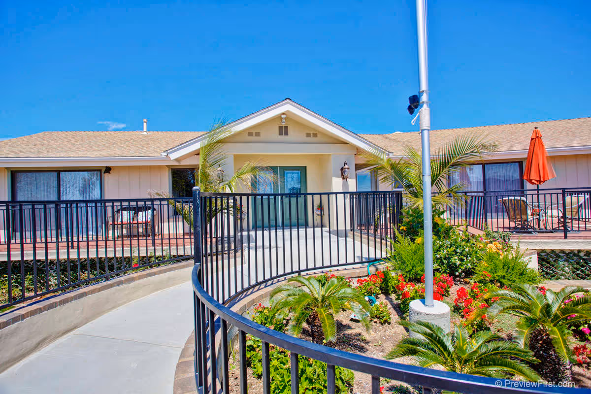 Exterior view of a single-story building with a beige facade and a brown shingled roof under a clear blue sky. There is a curved concrete pathway with black metal railings leading to a covered entrance with double doors. The landscaped garden in front features various green plants and colorful flowers. Patio furniture and a red umbrella are visible on the right side of the building.