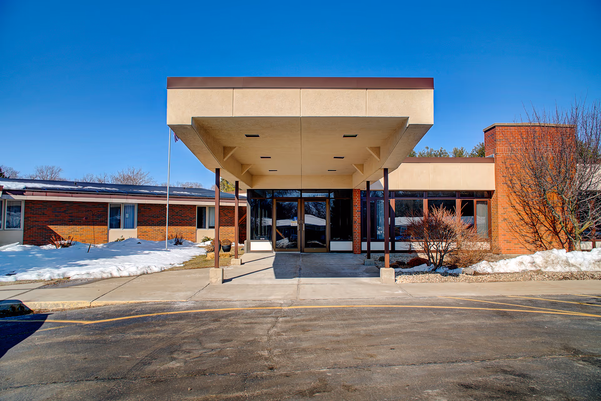 Front exterior view of Evansville Manor Skilled Nursing facility with a covered entrance, brick walls, large windows, and some snow on the ground under a clear blue sky.