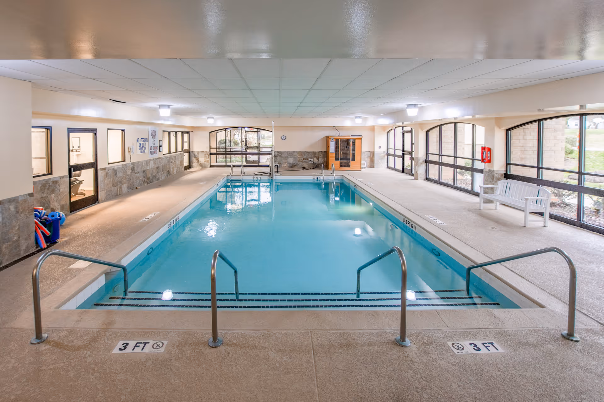 Indoor swimming pool with clear blue water, metal handrails, and steps leading into the pool. The pool area is surrounded by large windows letting in natural light, beige walls with stone accents, a white bench on the right side, and a sauna room at the far end. The ceiling has recessed lighting.