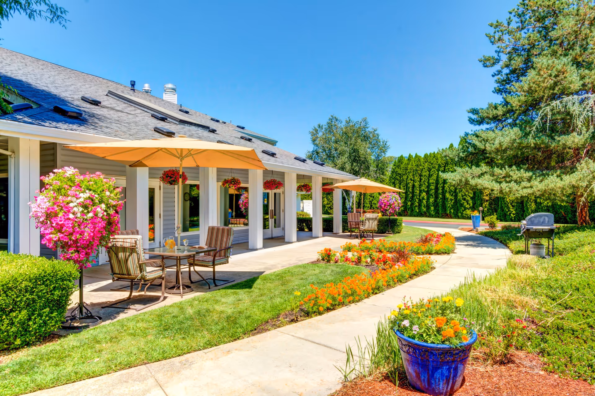 Outdoor patio area of The Suites Assisted Living and Memory Care facility featuring tables with umbrellas, chairs, hanging flower baskets, colorful flower beds, and a paved walkway surrounded by greenery and trees under a clear blue sky.