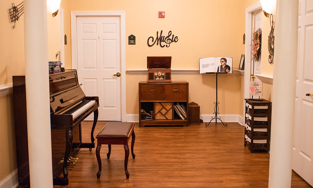 A warm interior common room featuring an upright piano with bench, a wooden music cabinet, a music stand, and a 'Music' wall sign.