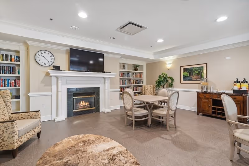 A cozy common area in a senior living facility featuring a fireplace with a TV mounted above it, built-in bookshelves filled with books on either side, a round table with four chairs, a floral armchair, a wooden sideboard with beverage dispensers, and a framed picture on the wall.