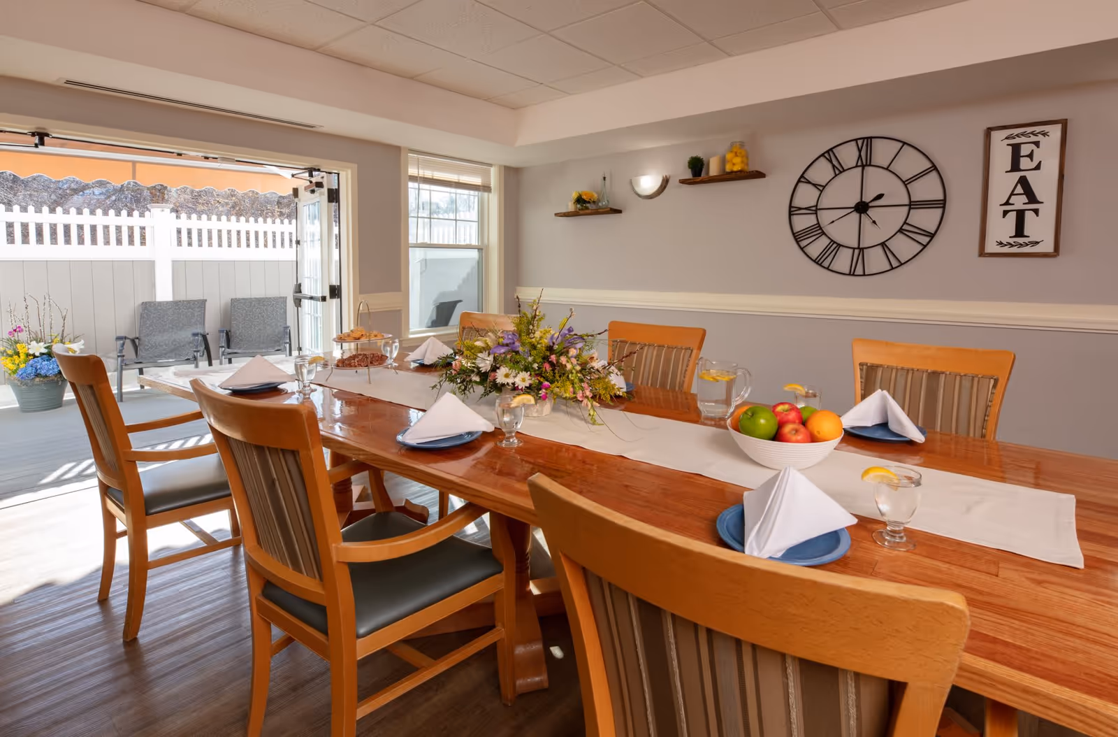A dining room with a long wooden table set for a meal, featuring folded white napkins on blue plates, glasses of water with lemon, a bowl of fruit, and a floral centerpiece. The room has light gray walls with a large clock and a sign that says 'EAT'. There are wooden chairs around the table and an open door leading to an outdoor patio with chairs and a white fence.