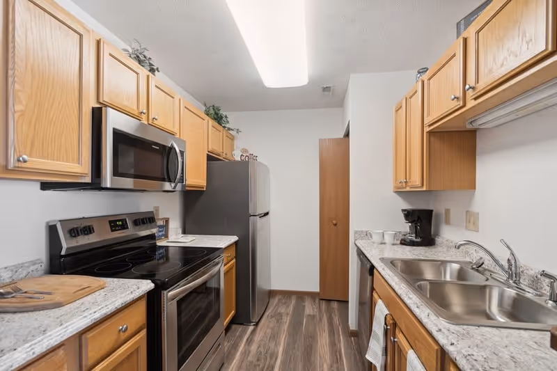 A modern kitchen with wooden cabinets, a stainless steel microwave and oven, a stainless steel refrigerator, a double sink, a coffee maker, and granite countertops. The floor is wood-patterned, and there is a fluorescent ceiling light.