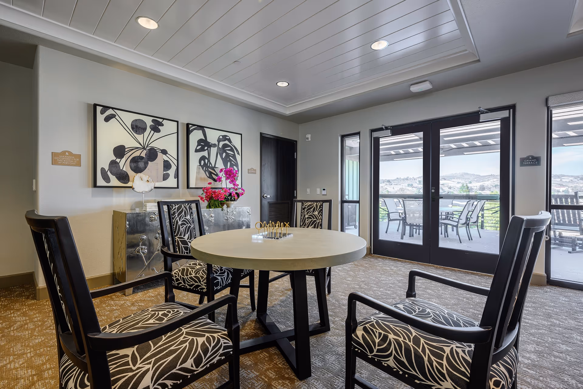Communal dining area with a round table and patterned chairs, wall artwork, and glass doors opening to an outdoor patio.