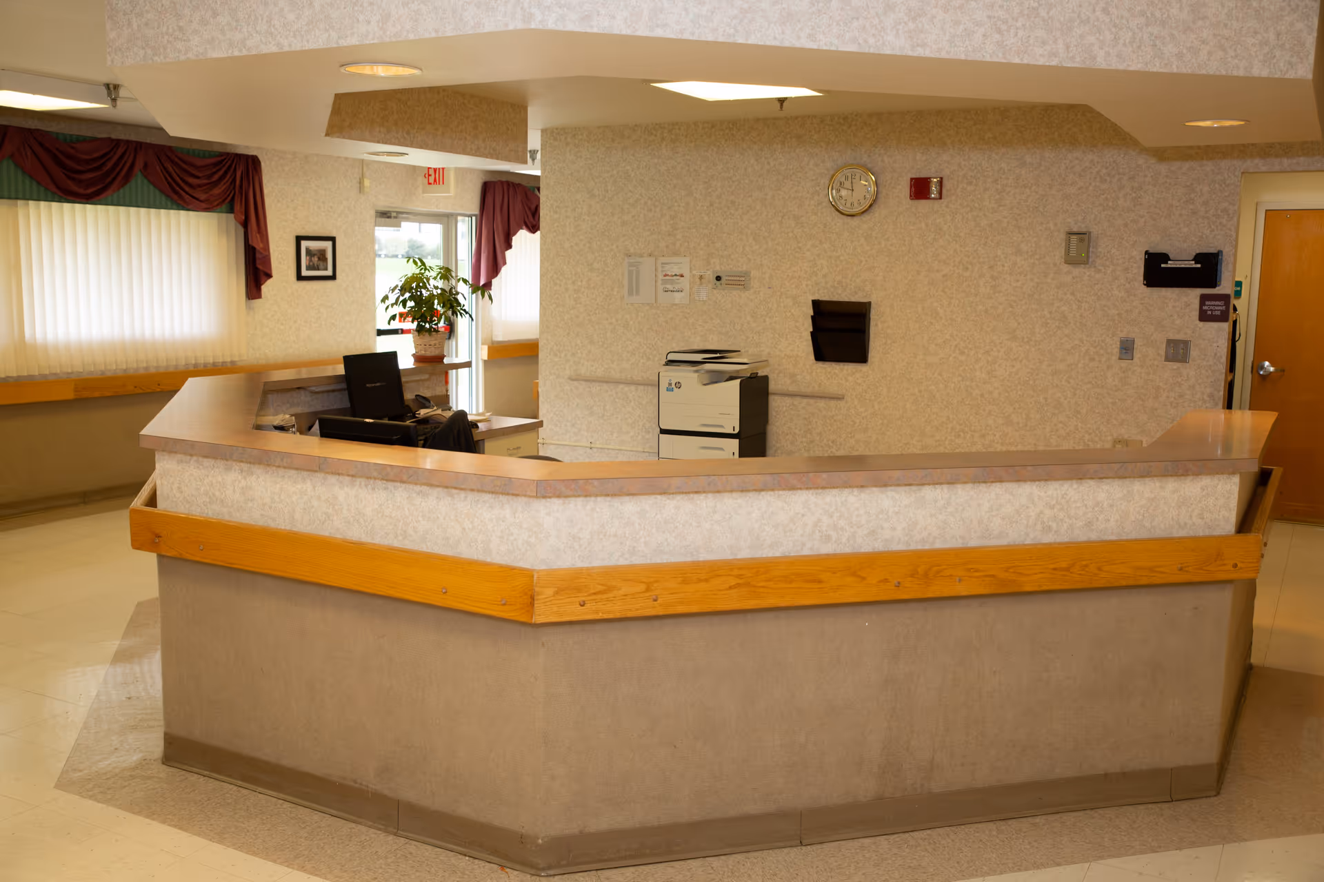 Empty reception desk area with a wraparound counter, office chair, printer, and wall clock inside a senior living facility.