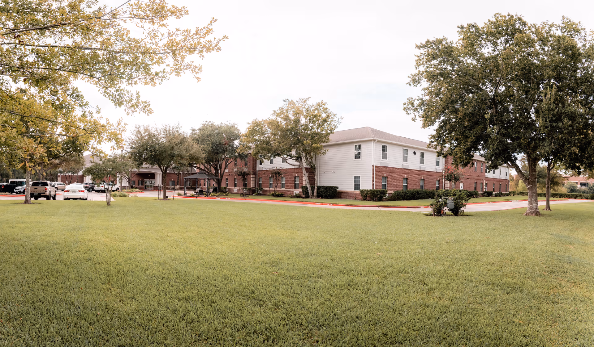 Exterior view of Greatwood at Sugar Land senior living facility showing a two-story building with red brick and white siding, surrounded by green grass, trees, and a parking area with several cars.
