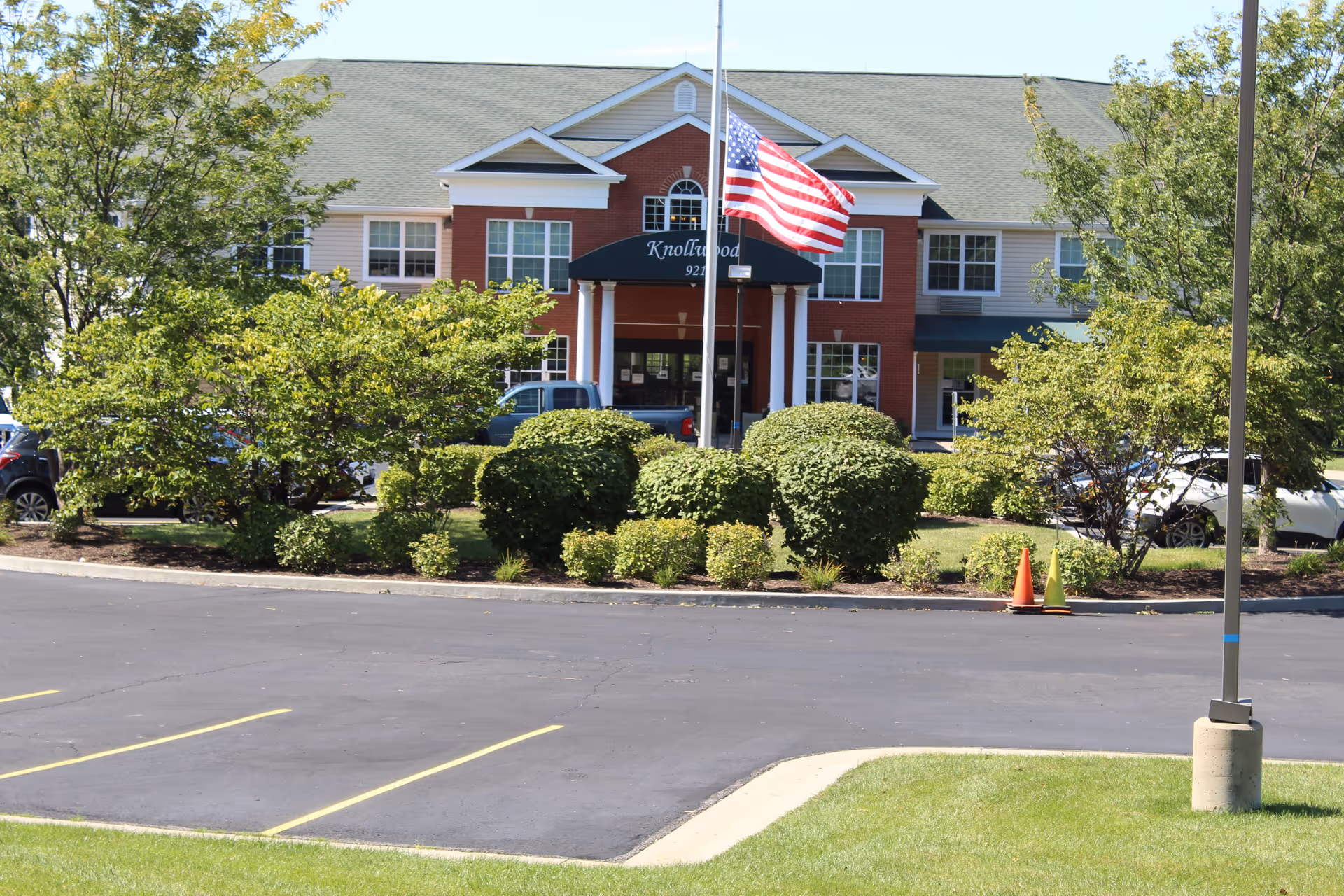 Front exterior view of Knollwood Retirement Community building with an American flag on a flagpole in front, surrounded by green bushes and trees, and a parking lot in the foreground.