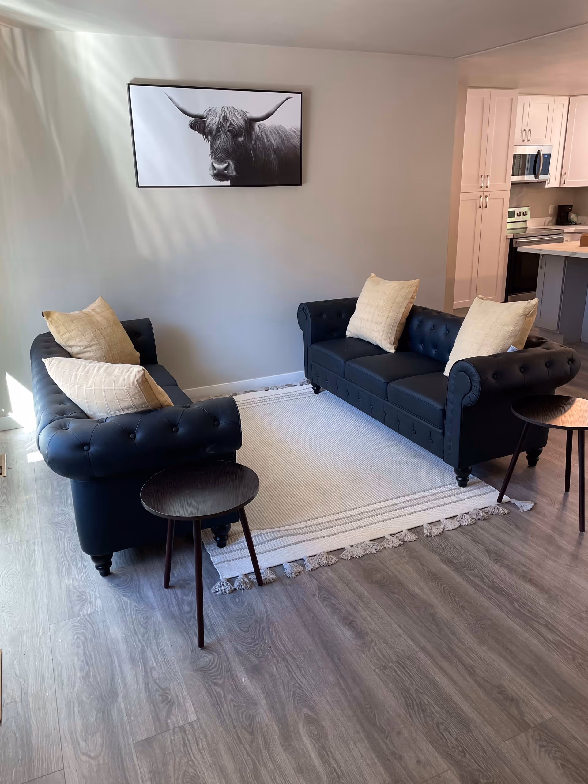 A modern living room with two black tufted sofas facing each other on a white rug, small round side tables, and a framed black-and-white cow photo on the wall with the kitchen visible in the background.