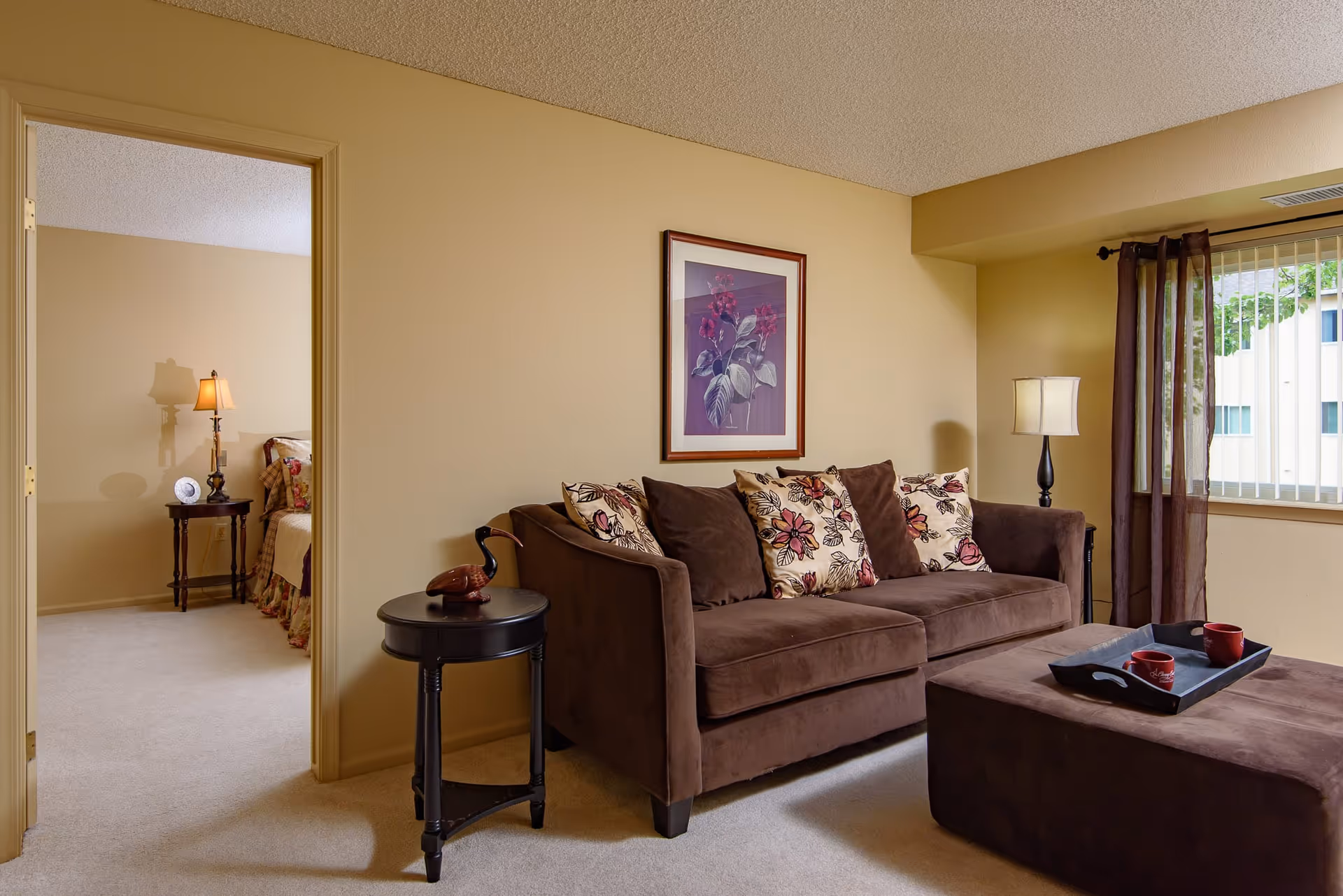 A cozy living room with a brown sofa adorned with floral and solid pillows, a matching ottoman with a tray holding two red mugs, a side table with a decorative bird sculpture, a floor lamp, and a window with sheer brown curtains. Through an open doorway, a bedroom with a bed, nightstand, and lamp is partially visible.