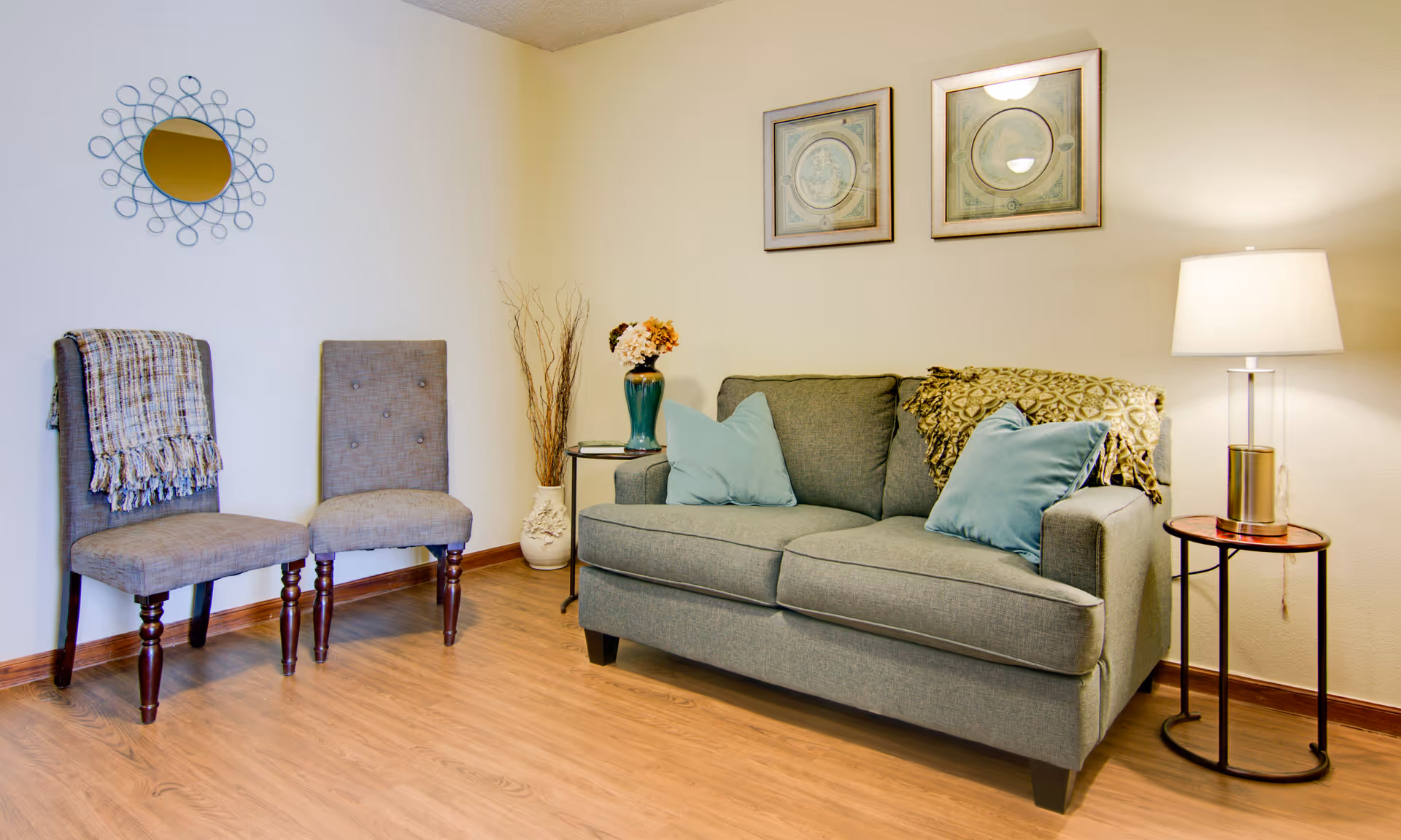 A cozy living room area with a gray sofa adorned with two blue pillows and a patterned throw blanket. Next to the sofa is a small round side table with a lamp. Two gray upholstered chairs are placed against the wall, one with a plaid throw draped over it. The walls are decorated with two framed pictures and a decorative round mirror. The floor is wooden.