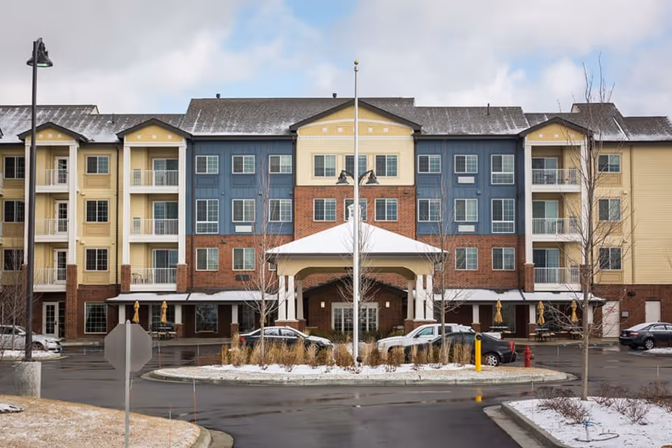 Front exterior view of a multi-story senior living facility building with a covered entrance, parked cars, and a roundabout with landscaping in front. The building has a mix of yellow, blue, and brick facade with balconies and windows.