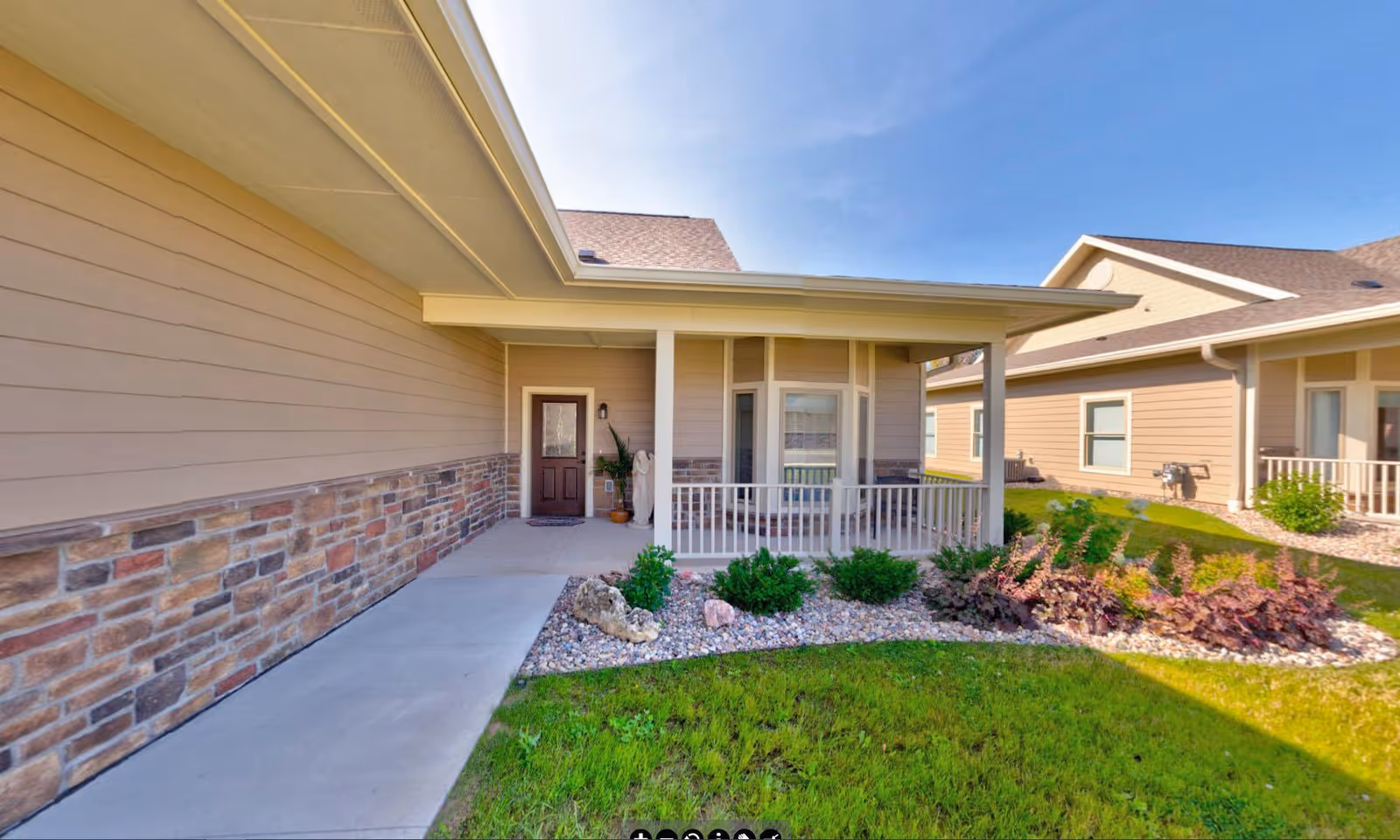 Exterior view of a single-story building with beige siding and stone accents, featuring a covered porch with white railing, a brown door, and a landscaped garden with green grass, bushes, and rocks under a clear blue sky.