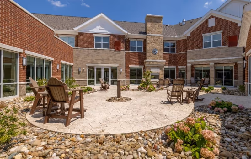 Outdoor courtyard area of a senior living facility with stone-paved circular seating area surrounded by rocks and plants. Several wooden chairs and tables are arranged around the courtyard. The building is two stories with brick and stone exterior walls, multiple windows, and a central chimney with a clock mounted on it under a clear blue sky.