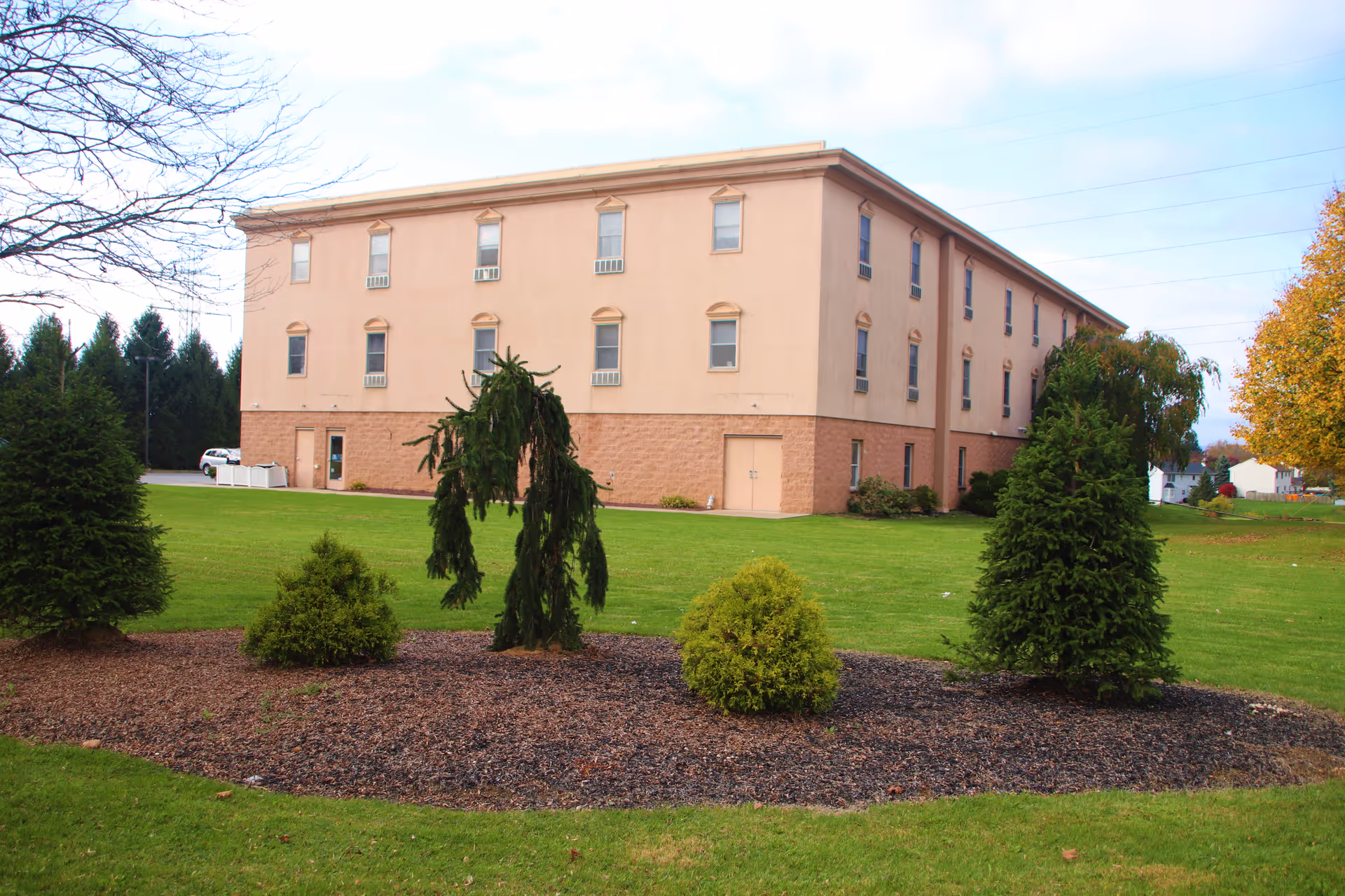 A three-story beige building with multiple windows, surrounded by a well-maintained lawn and landscaped area with small evergreen trees and shrubs. The sky is partly cloudy and there are some trees in the background.