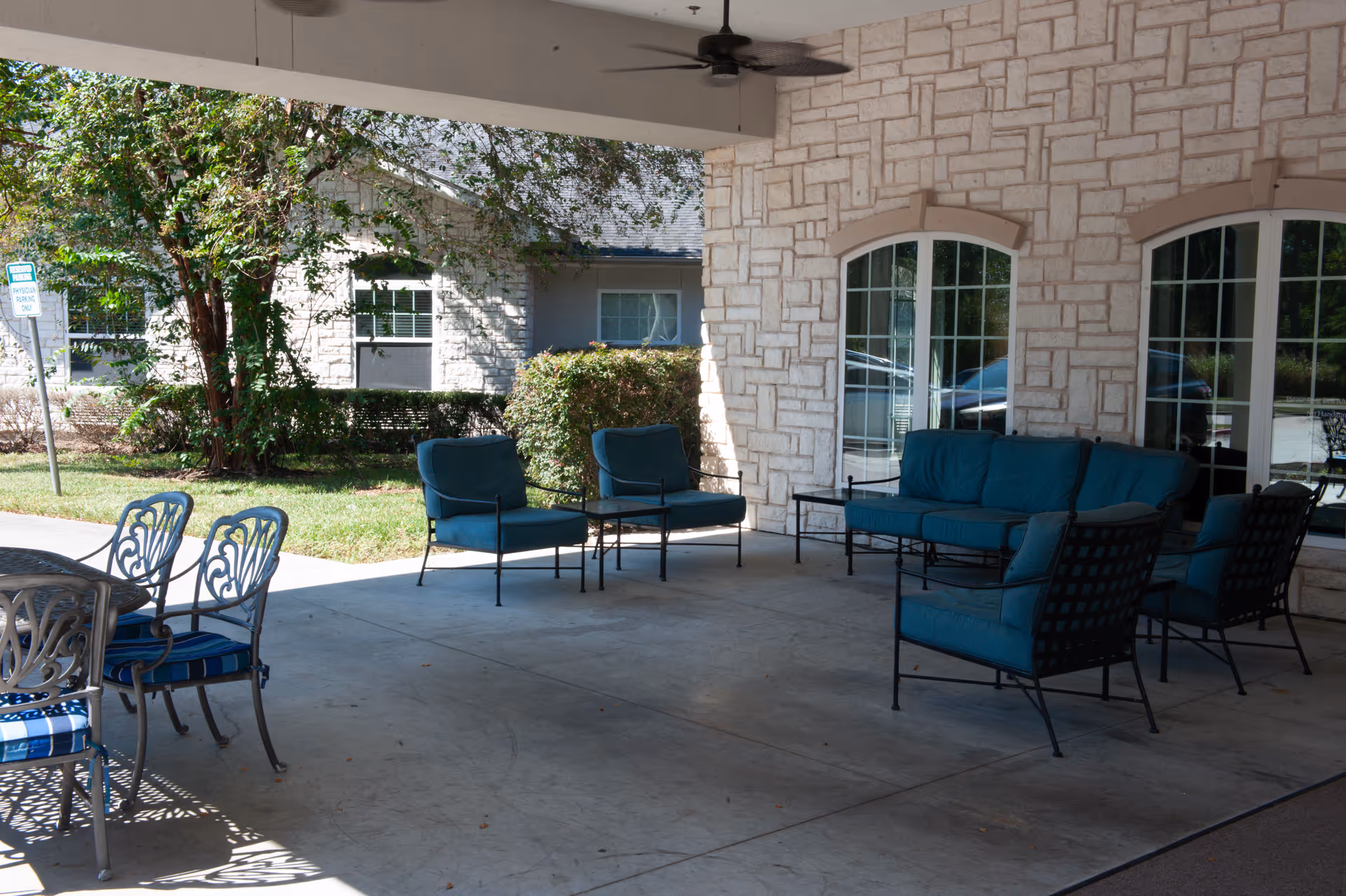 Covered outdoor patio area with cushioned metal chairs and a sofa arranged around small tables, adjacent to a stone building with large windows. Greenery and another building are visible in the background.